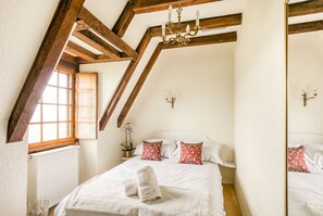A bedroom with a sloped ceiling, wooden beams, a bed with white linens, red floral pillows, a chandelier, and a window with a view. - Belle Vue - Four Bedroom Villa, Sleeps 8 (Castelnaud-la-Chapelle)