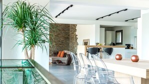 Un espace de repas moderne avec une table en verre et des chaises transparentes, un mur d’accent en pierre et une cuisine en arrière-plan.