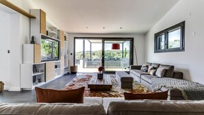Un salon moderne avec un canapé, une table basse et un téléviseur. Une grande fenêtre offre une vue sur la verdure à l’extérieur.