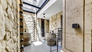 Un couloir aux murs de pierre, avec deux grandes barriques, un étagère en métal et une porte.