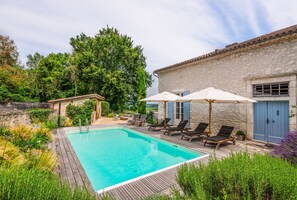 A swimming pool with lounge chairs and umbrellas, surrounded by greenery and a stone building.