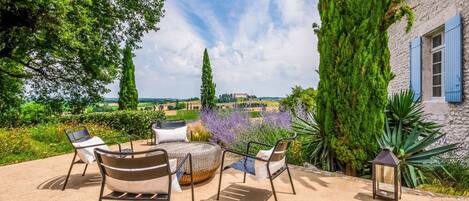 An outdoor seating area with a round table and chairs, surrounded by greenery and a view of the countryside.