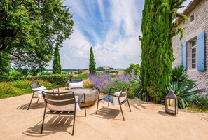 An outdoor seating area with a round table and chairs, surrounded by greenery and a view of the countryside.