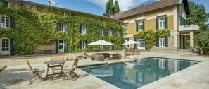A pool area with outdoor furniture, a clear pool, and a building with green shutters and ivy-covered walls.