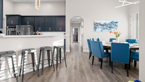 A modern kitchen with dark cabinets and stainless steel appliances, a dining area with blue chairs and a wooden table, and a hallway leading to another room. - BILTMORE BLISS→Rooftop w/ Mountain/City View, Pool (Phoenix)