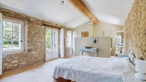A bedroom with a stone wall, wooden beams, a bed with a white bedspread, a small desk, and a mirror. - Mas Des Collines - Six Bedroom Villa, Sleeps 12 (Saignon)