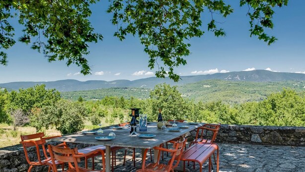 An outdoor dining area with a stone table and orange chairs, set against a backdrop of lush green hills and mountains. - Mas Des Collines - Six Bedroom Villa, Sleeps 12 (Saignon)