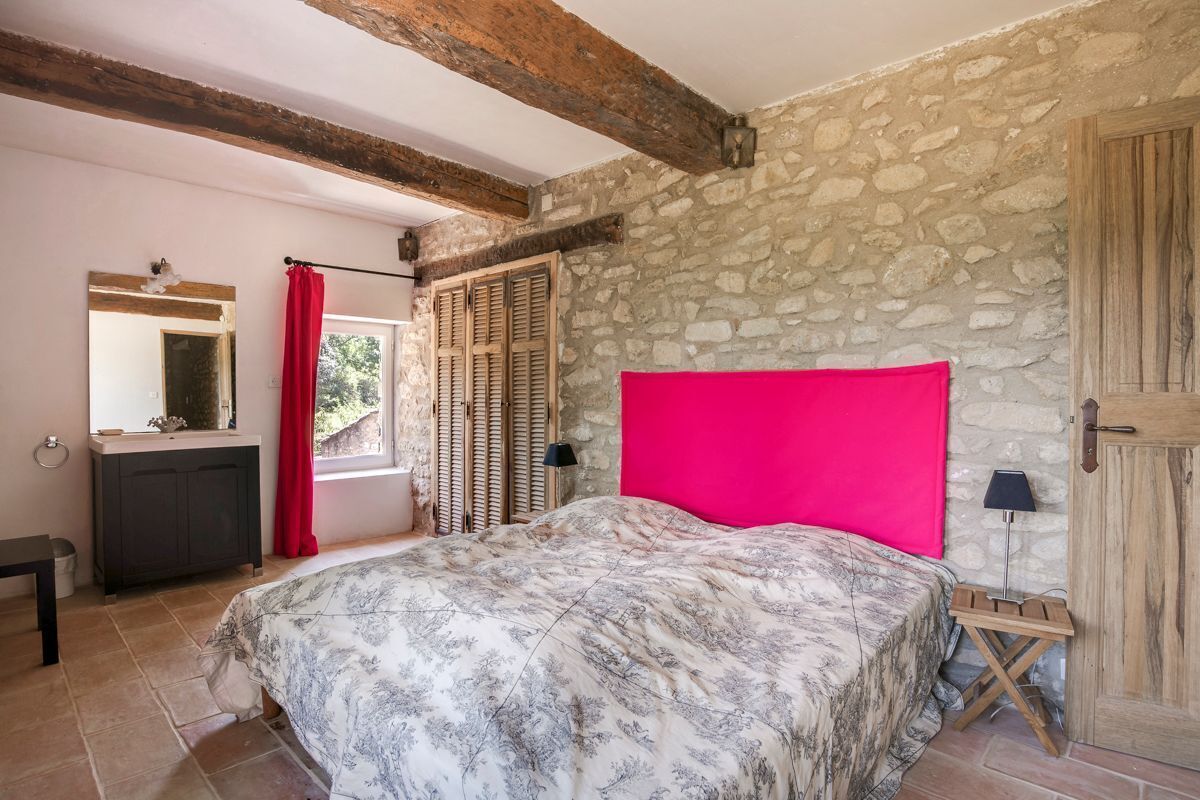 Quarto com parede de pedra, cama com colcha estampada, cabeceira de madeira, mesa de cabeceira, penteadeira, espelho e janela com persianas.