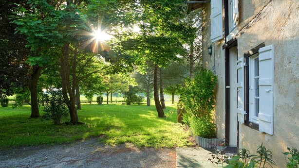 A house with a gravel driveway leading to a wooden door, surrounded by lush greenery and trees. - Cottage Des Cathares (Plaigne)