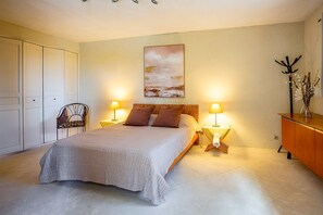 A bedroom with a bed, two bedside lamps, a chair, a desk, a cabinet, and a painting on the wall.