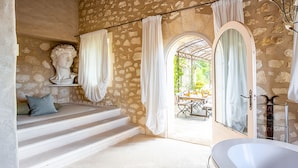 Ein Badezimmer mit Steinwänden, einer Badewanne, einer Statue und Blick auf eine Terrasse.