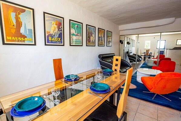A dining area with a long table, chairs, and a glass-top table. There are framed pictures on the wall.