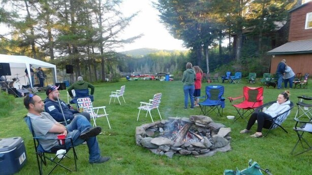 Eine gesellige Runde im Freien, Menschen sitzen auf Rasensesseln um ein Feuerkörbchen, im Hintergrund ein Zelt und verschiedene Gegenstände.