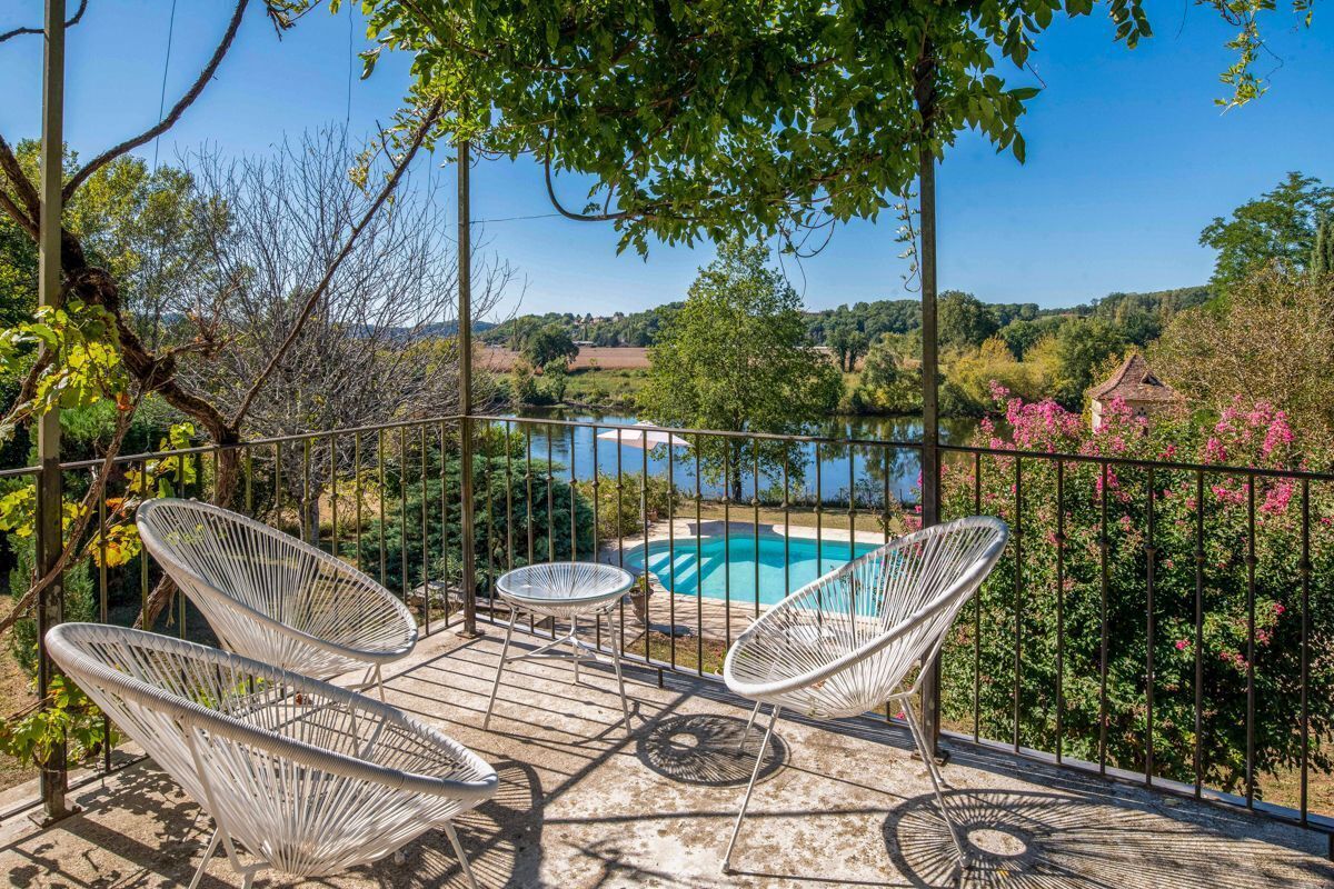 Une terrasse dotée de chaises en osier blanc et d’une petite table, offrant une vue sur une piscine et une végétation luxuriante.