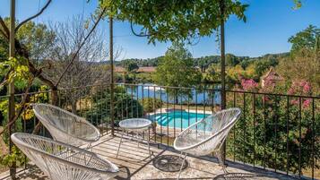 Une terrasse dotée de chaises en osier blanc et d’une petite table, offrant une vue sur une piscine et une végétation luxuriante.