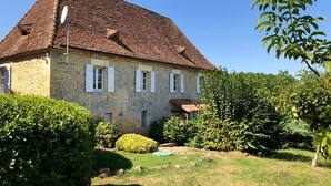 Ein Haus aus Stein mit Ziegeldach, weißen Fensterläden und einem Garten mit verschiedenen Pflanzen und einem kleinen Teich.