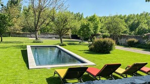 Une piscine rectangulaire bordée de pierres, entourée d’une pelouse bien entretenue. On y trouve trois chaises longues avec des coussins rouges et une table jaune.