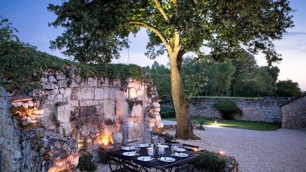 Un espace extérieur aménagé pour les repas, avec un mur en pierre, un arbre et un sol recouvert de gravier.