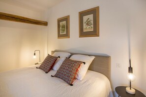 Ein ordentlich bezogenes Bett mit gemusterten Kissen, einem Kopfteil und zwei gerahmten Pflanzenbildern an der Wand.