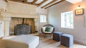Une pièce avec une cheminée, un lit, deux poufs, un fauteuil en osier et une fenêtre avec un tableau accroché au mur.