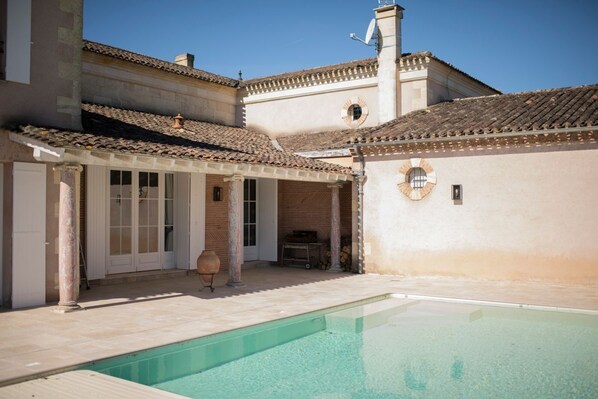 Un espace piscine doté d’une terrasse carrelée, d’un coin salon abrité et d’un bâtiment avec un toit et des colonnes recouverts de carreaux.