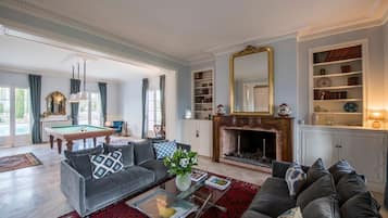 Een ruime woonkamer met een pooltafel, banken, een open haard en een boekenkast.