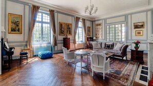 Una habitación amplia con un piano, un reposapiés azul, una silla blanca, un sofá y una araña.