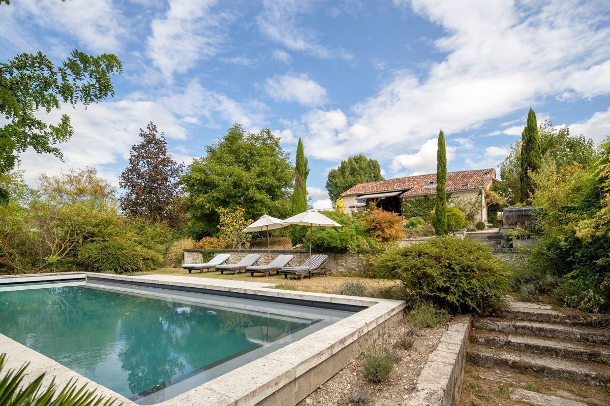 Une piscine entourée d’arbres, agrémentée de chaises longues et de parasols, et à laquelle on accède par un escalier en pierre.