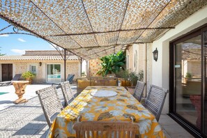 Une terrasse extérieure avec une table dressée pour le repas, entourée de chaises en osier et d’une nappe à motifs.
