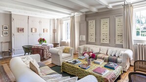 A cozy living room with a sofa, armchairs, a coffee table, and a dining table. There are framed pictures on the walls and a window with curtains.