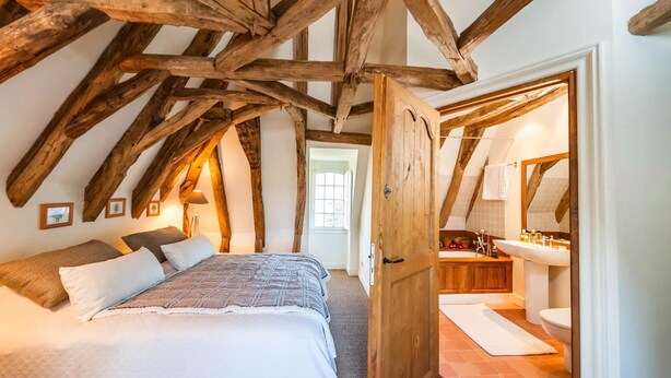 A bedroom with a sloped ceiling, wooden beams, a bed with white bedding, and a view into a bathroom with a sink and mirror. - Chartreuse Des Roses (Pinsac)