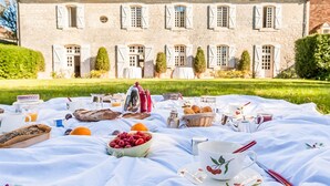 A picnic setup with food and drinks on a blanket in front of a large stone building with multiple windows and a tiled roof. - Chartreuse Des Roses (Pinsac)