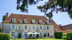 A stone building with a symmetrical facade, multiple windows with shutters, and a well-maintained lawn with outdoor seating. - Chartreuse Des Roses (Pinsac)