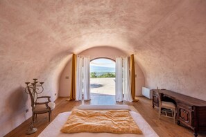 Une chambre confortable avec un grand lit, un bureau et une vue sur l’extérieur, visible à travers une porte en arc de cercle.
