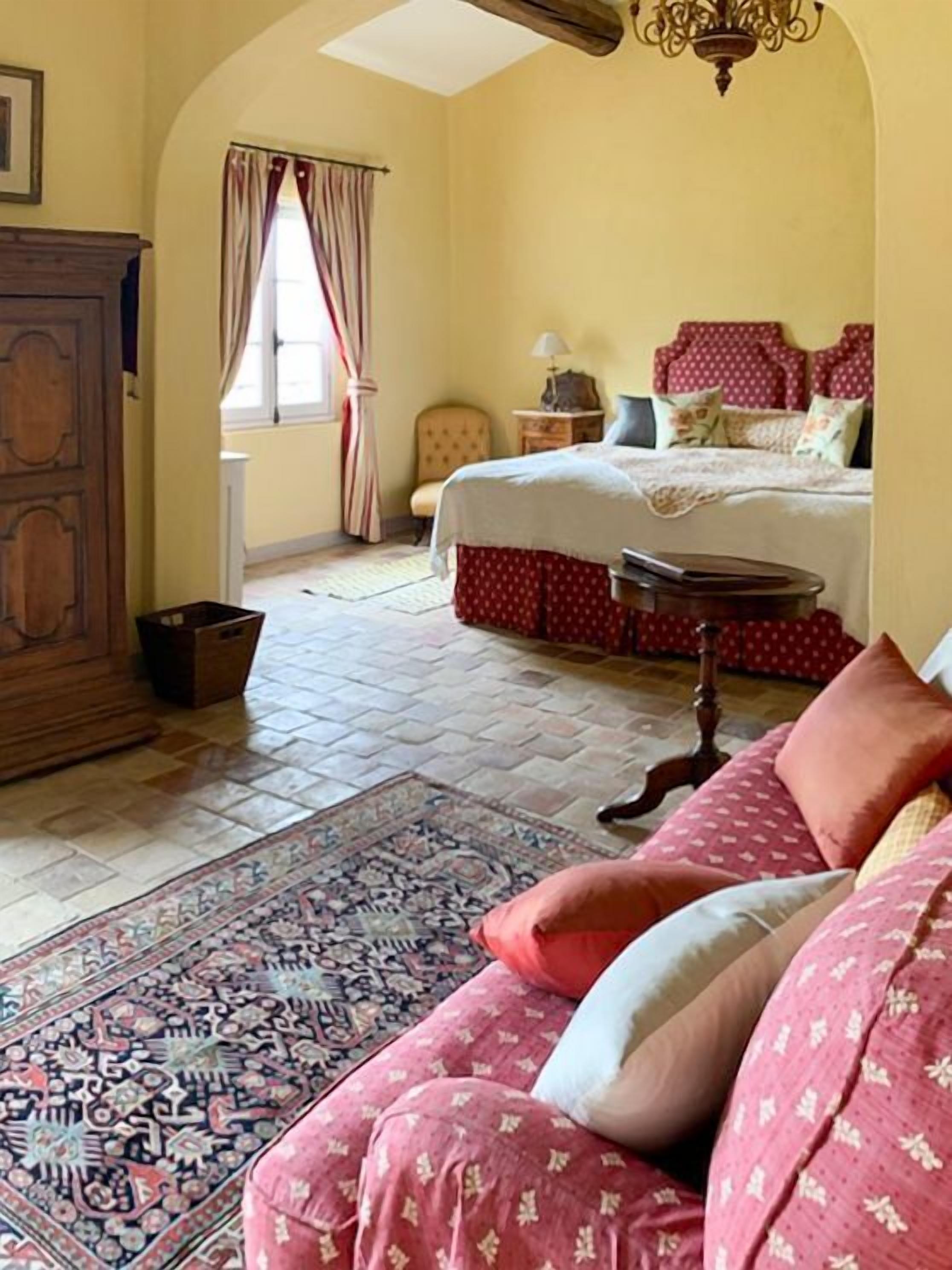 A bedroom with a bed, a wooden dresser, a patterned rug, and a chandelier.