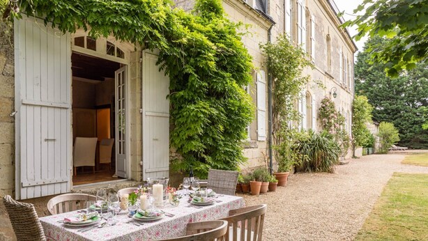 Un espace de restauration en plein air avec une table dressée pour deux personnes, entouré de plantes en pot et bordé d’un bâtiment à la façade en pierre.