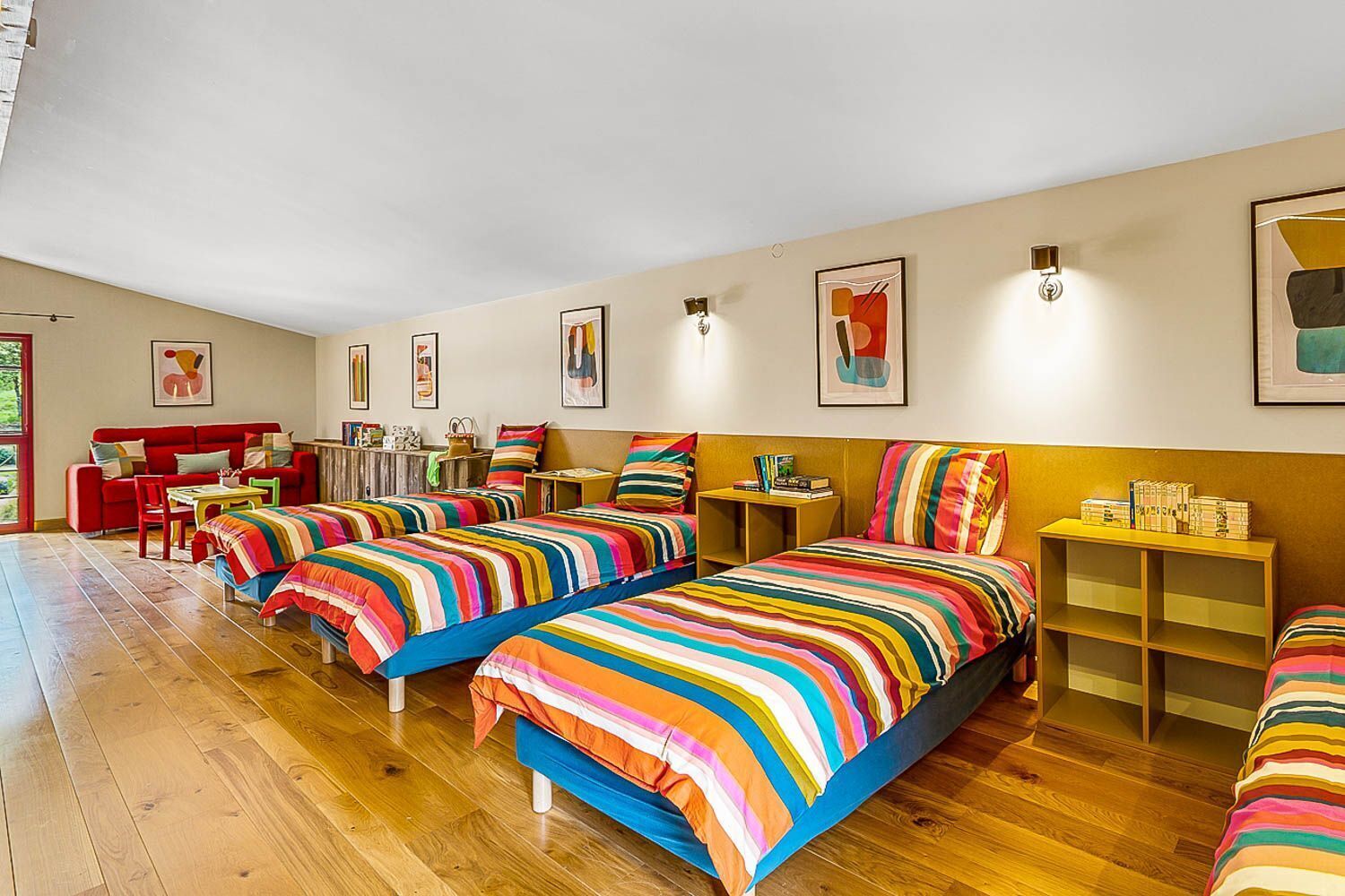 Een kamer met drie bedden, een rode bank, een tafeltje en een boekenkast.
