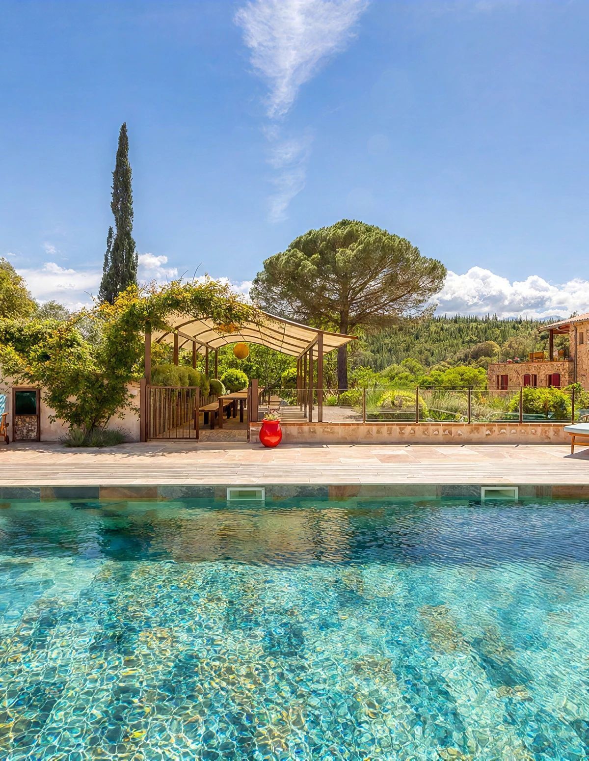 Une piscine aux eaux cristallines, agrémentée d’une pergola et d’un espace détente, le tout entouré d’arbres et d’un bâtiment.