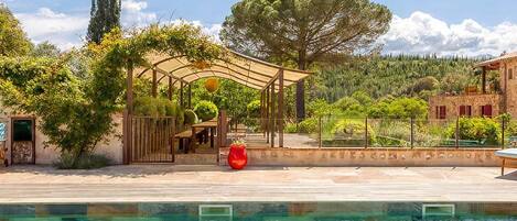 A clear swimming pool with a pergola and seating area, surrounded by trees and a building.
