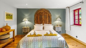 A bedroom with a large bed, two bedside tables, a wooden nightstand, a framed picture on the wall, and a window with a red frame.