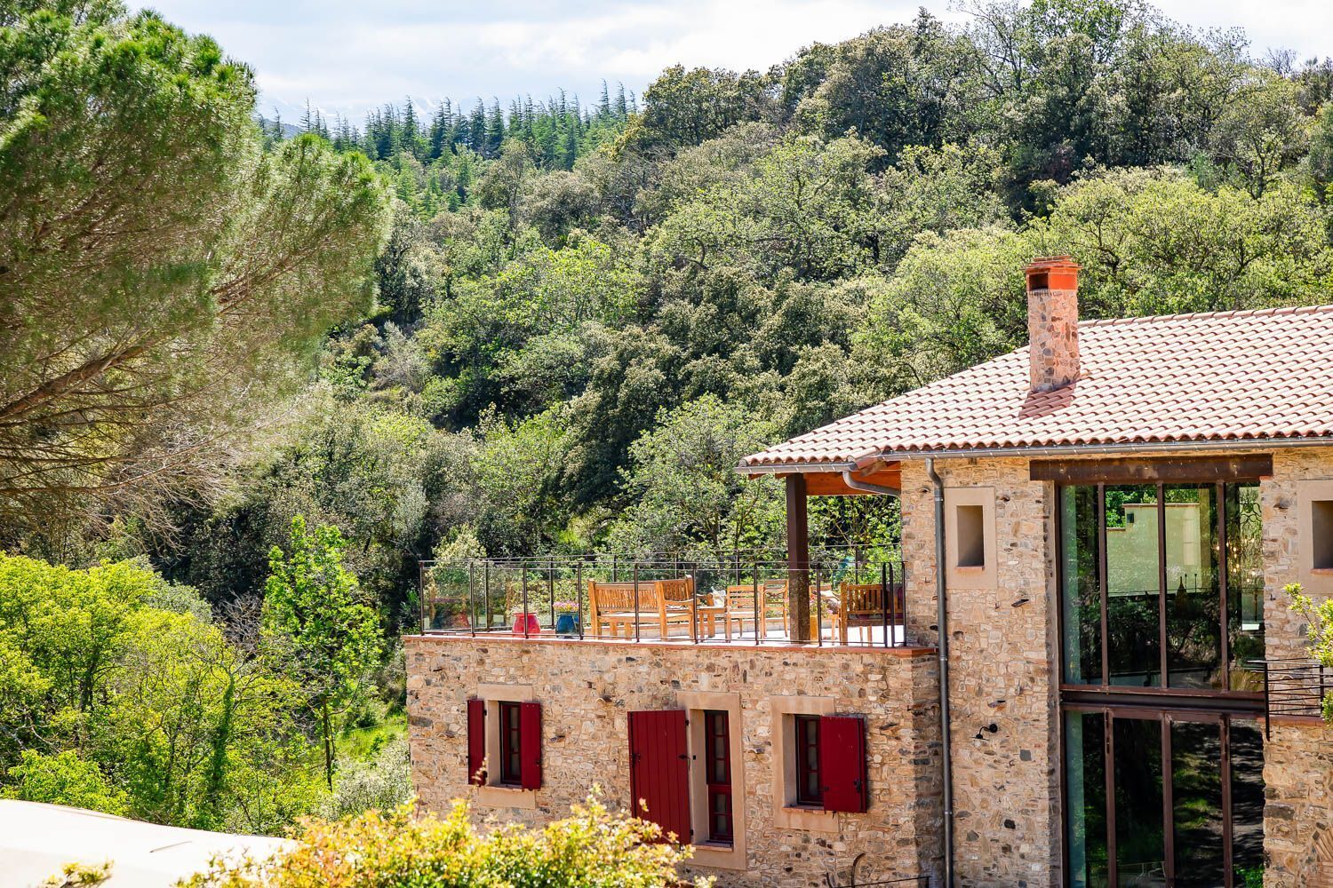 A stone building with a tiled roof, red shutters, and a balcony overlooking a forested hillside.