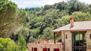 A stone building with a tiled roof, red shutters, and a balcony overlooking a forested hillside. - Domaine Les Pyrenees (Castelnou)