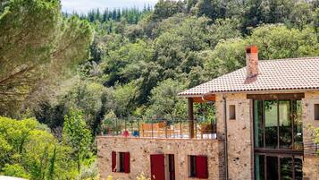 A stone building with a tiled roof, red shutters, and a balcony overlooking a forested hillside.
