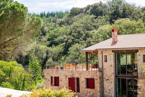 A stone building with a tiled roof, red shutters, and a balcony overlooking a forested hillside. - Domaine Les Pyrenees (Castelnou)
