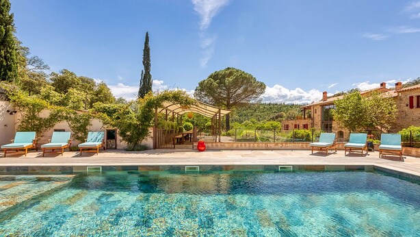 A clear swimming pool with lounge chairs and a pergola, surrounded by trees and a stone building.
