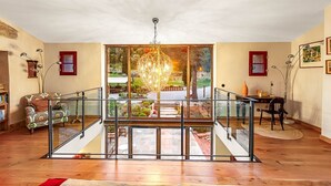 A spacious living area with a glass staircase, a chandelier, and a view of a garden through large windows.