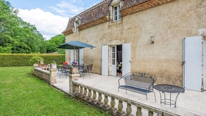 Un bâtiment en pierre avec un toit en tuiles, un patio couvert avec des meubles d’extérieur et une haie en arrière-plan.