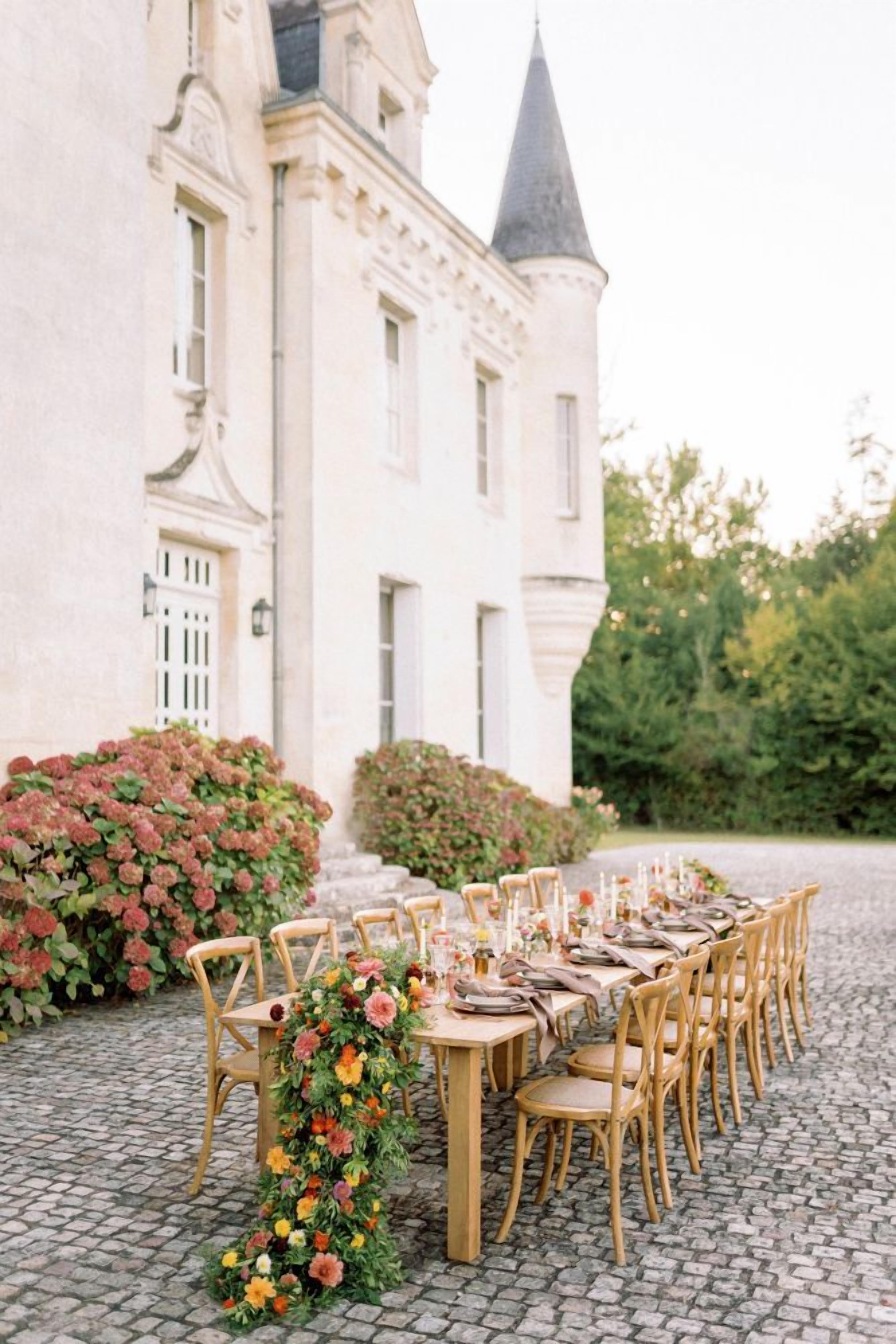 Een buitenterras met een lange tafel en stoelen, omgeven door weelderige begroeiing en een historisch gebouw op de achtergrond.