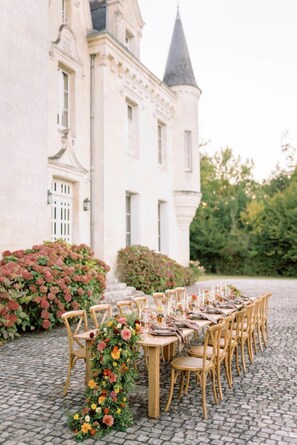Un espace de restauration en plein air avec une longue table et des chaises, entouré d’une végétation luxuriante et avec un bâtiment historique en arrière-plan.