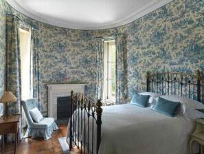 Une chambre à coucher avec un grand lit, une chaise, une cheminée et une fenêtre avec des rideaux.